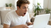 Young Caucasian man in white t-shirt looking shocked and surprised while using laptop computer at home, expressing amazement at screen content.