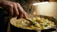 Professional chef hand stirring creamy potato gnocchi with herbs in cast iron skillet. Culinary cooking process with steam and warm kitchen lighting.