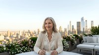 Professional mature woman with gray hair in white blazer sitting on urban rooftop terrace with city skyline background during golden hour sunset.