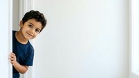 Young boy with dark curly hair and medium skin tone peeking around white wall corner, wearing navy blue shirt, smiling at camera. Suitable for family, education, and lifestyle designs.