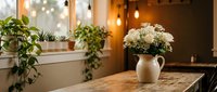Cozy kitchen interior with white chrysanthemum flowers in ceramic pitcher on wooden table, houseplants on windowsill, warm ambient lighting creating homey atmosphere.