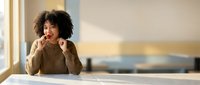 Young woman with curly dark hair and medium skin tone sitting at white desk, wearing beige sweater, resting chin on hands with thoughtful expression, natural window lighting, blurred office background.
