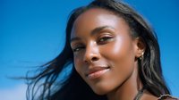 Young African American woman with glowing skin and radiant smile against vibrant blue sky background, hair flowing in breeze, expressing joy and confidence in natural outdoor lighting.