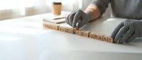 Professional businessman arranging wooden letter blocks spelling BUSINESS GRANT on white desk wearing black gloves in modern office environment.