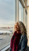 Happy mature African American woman with gray curly hair smiling at airport terminal window overlooking planes on tarmac during golden hour travel moment.