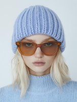 Young Caucasian woman with blonde hair wearing light blue knitted beanie hat, amber sunglasses and matching sweater against white background, fashion portrait.