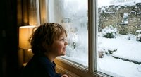 Young boy with curly hair gazing thoughtfully through window at snowy winter landscape, warm indoor lighting creating cozy atmosphere for seasonal content.