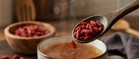 Wooden spoon adding dried goji berries to steaming hot chocolate drink in ceramic mug. Superfood ingredient for healthy beverages and wellness recipes.