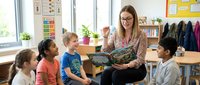 Adult female teacher with brown hair and glasses reading a storybook to diverse elementary school children sitting on floor in bright classroom with educational posters and shelves.