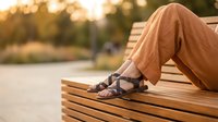 Woman in beige pants and brown sandals relaxing on wooden park bench during golden hour sunset, perfect for lifestyle and leisure concepts.