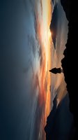 Silhouetted figure standing on rocky cliff edge overlooking dramatic golden sunset sky with vibrant orange and amber clouds reflecting on distant horizon, contemplative outdoor scene.