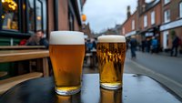 Two fresh pints of golden beer on outdoor table at busy street pub with blurred pedestrians and historic brick buildings in background during evening hours.
