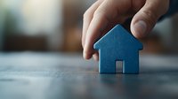 Small blue wooden house model held by fingers against blurred background, representing real estate, property investment and home ownership concept.