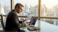 Professional businessman working on laptop in modern office with city skyline view during golden hour, corporate executive at desk with coffee cup.