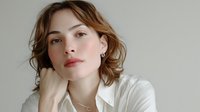 Young woman with wavy brown hair in white shirt against neutral background, hand resting under chin in contemplative pose.
