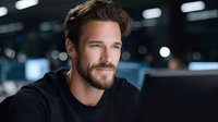 Confident young Caucasian businessman with beard smiling in modern office environment. Professional male entrepreneur working late at corporate workplace.