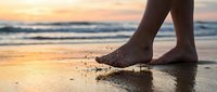 Bare feet walking on wet sand at sunset beach, water splashing, warm golden light reflecting on shore, summer vacation freedom concept.