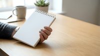 Woman's hand holding open blank spiral notebook at wooden desk with coffee cup and succulent plant, workspace organization and planning concept.