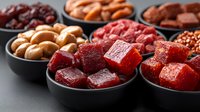 Fresh fruit and berry cubes in black bowls on dark background, healthy snacks assortment with dried cranberries, nuts and candied fruits.