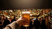 Cropped hand holding beer glass at festive outdoor gathering with golden string lights and crowd in background, suitable for holiday marketing and beverage advertising.