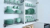 Organized kitchen cabinet with mint green ceramic dishes, bowls, cups, and plates neatly arranged on chrome wire racks in white cupboards.