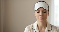 Young woman wearing white sleep mask relaxing with closed eyes in soft natural light for wellness and self-care routine.