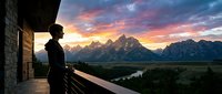 Woman in dark clothing standing on modern balcony overlooking dramatic mountain range at sunset with river valley below, scenic landscape photography for travel and lifestyle use.