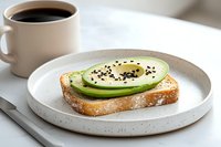 Fresh avocado toast with sliced green avocado, black sesame seeds and soft boiled egg on sourdough bread, served with black coffee on ceramic plate.