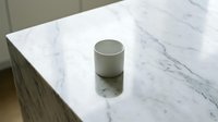 Cylindrical metallic container on white marble surface with veining, reflecting light, suitable for product mockups and luxury branding designs.