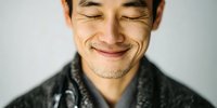 Close-up portrait of Asian man with eyes closed and gentle smile, wearing dark textured coat against neutral background.