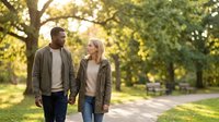 Happy interracial couple walking hand in hand through sunny park during golden hour, enjoying romantic outdoor date together in natural green setting.