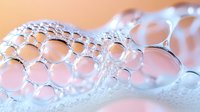 Macro photography of soap bubbles forming delicate foam pattern against pink background, showing transparent spherical structures with light reflections and surface tension.