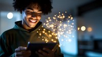 Young African American woman with natural curly hair smiling while using tablet device with magical glowing sparkles and light effects emerging from screen in dark room.