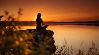 Woman with long dark hair, sitting on a rock overlooking a calm lake at golden hour sunset, silhouetted against warm orange and amber sky, peaceful meditation and contemplation scene.