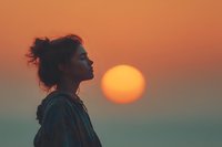 Young woman in profile silhouette against vibrant orange sunset sky, peaceful moment of contemplation and mindfulness during golden hour.