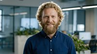 Smiling bearded Caucasian businessman with curly hair in navy shirt standing confidently in modern office workspace for corporate communications.
