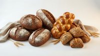 Assorted artisan breads and baked goods including whole grain loaves, pretzel rolls, and seeded rolls arranged with wheat stalks on neutral background for bakery and food styling.