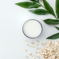 Glass of oat milk with scattered oats and green leaves on white background, flat lay composition for healthy lifestyle and plant-based diet concept.