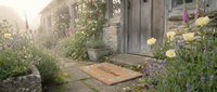Rustic cottage entrance with welcome mat, blooming lavender and wildflowers, weathered stone walls and wooden door in soft morning light.