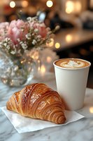 Fresh buttery croissant and latte art coffee on marble table with pink roses and baby breath flowers in soft bokeh background. Cozy breakfast atmosphere.