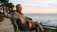 Senior man with gray hair and Caucasian ethnicity sitting on bench by coastal path at sunset, wearing casual jacket, gazing toward ocean horizon. Suitable for lifestyle, retirement, wellness, and travel concepts.