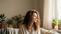 Young Caucasian woman with curly hair smiling and gazing out window at home, surrounded by green plants in cozy living room.