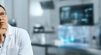 Mature male scientist or researcher wearing white lab coat and glasses, standing in modern laboratory environment with blurred medical equipment and monitors in background, contemplative expression.