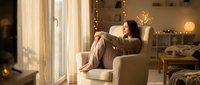 Young woman relaxing in cozy armchair by window with warm lighting and fairy lights creating peaceful home atmosphere for wellness content.