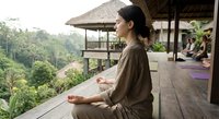 Young Asian woman practicing meditation in lotus pose on wooden deck at tropical wellness retreat surrounded by lush green forest and traditional architecture.