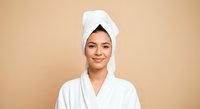 Woman with wrapped hair towel wearing white bathrobe after shower or spa treatment against beige background for wellness and beauty content.