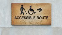 Accessible route sign with braille text and disability symbols showing walking person with cane and wheelchair user with directional arrow on bronze plaque.