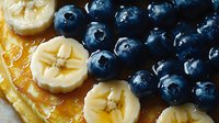 Fresh blueberries and sliced banana on golden pancakes drizzled with maple syrup, close up view showing texture and details of breakfast dish.