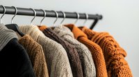 Cozy knitted sweaters in earth tones hanging on clothing rack against white wall. Warm autumn and winter wardrobe collection featuring beige, brown, and orange colors.
