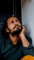 Young Hispanic man with beard and long hair looking up thoughtfully while leaning against white wall, wearing dark green shirt and yellow t-shirt underneath.